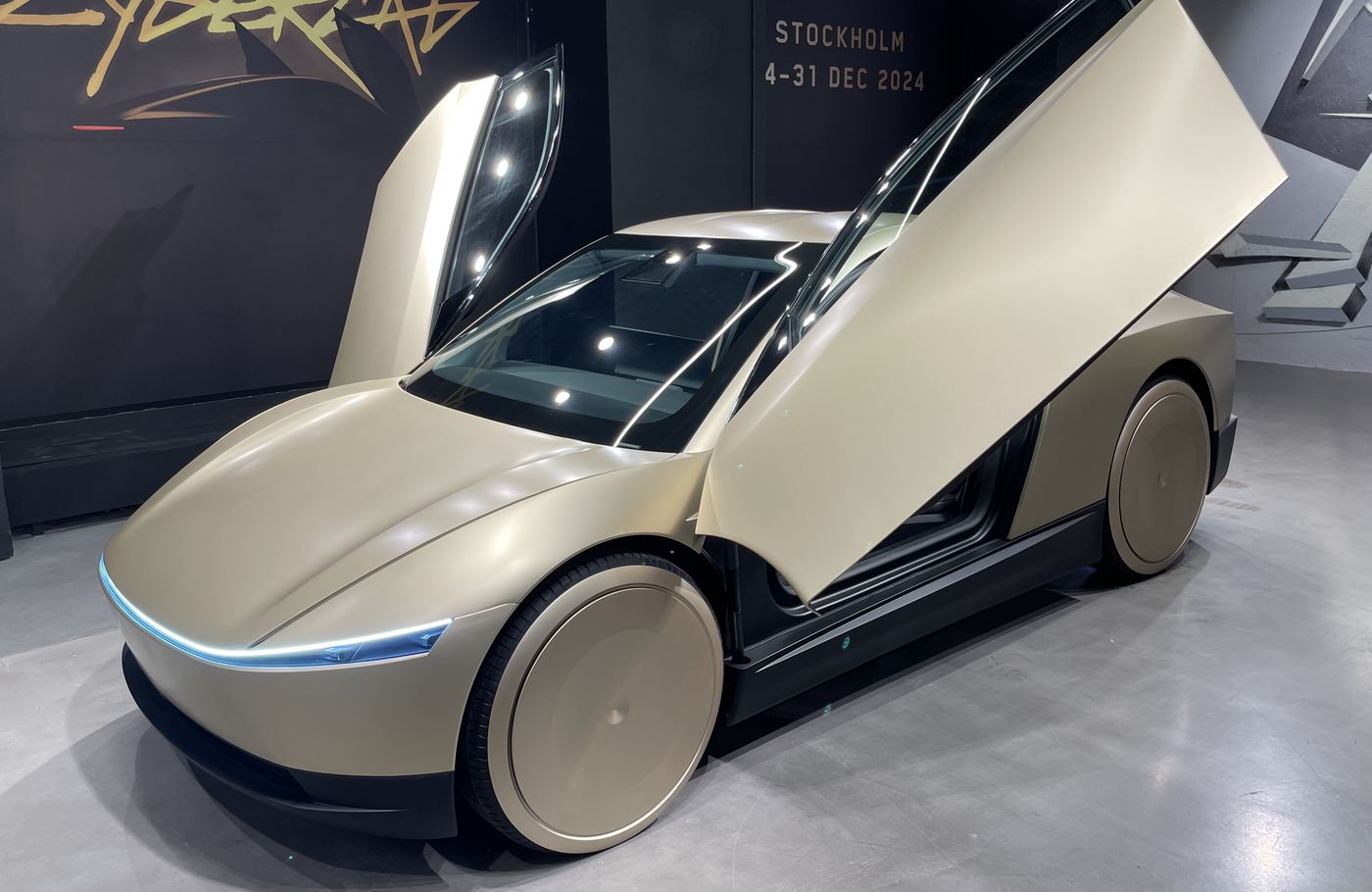 Front three-quarter view of a champagne Cybercab in a dark showroom, both upward-opening doors raised and a thin blue light strip across its nose.