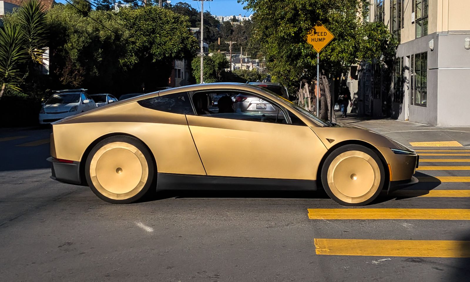 A champagne-gold two-seat Cybercab rolls along a sunny tree-lined city street, seen from the side as it crosses a yellow-striped crosswalk.