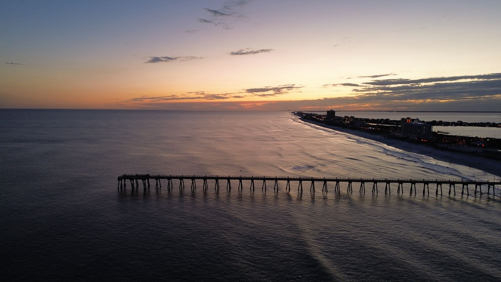 Aerial view of the Pensacola Beach Gulf Pier at sunset, the pier stretching into calm water under an orange and violet sky.
